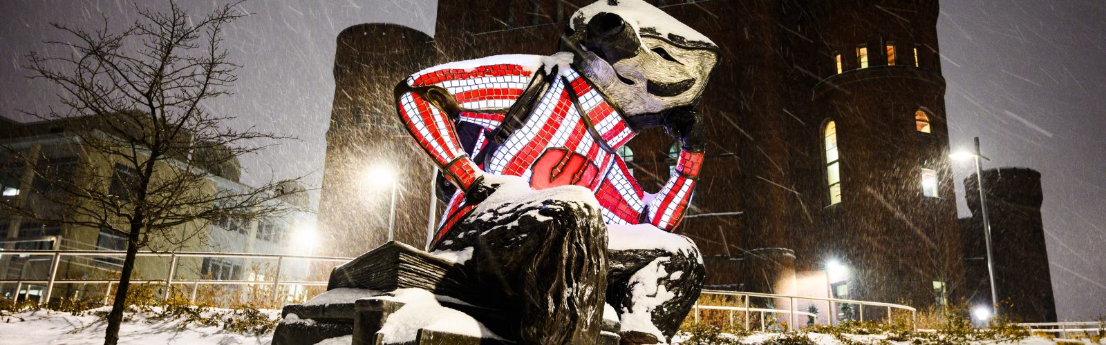 Bucky badger statue at Alumni Park with Red gym, a tall turreted building, in the winter