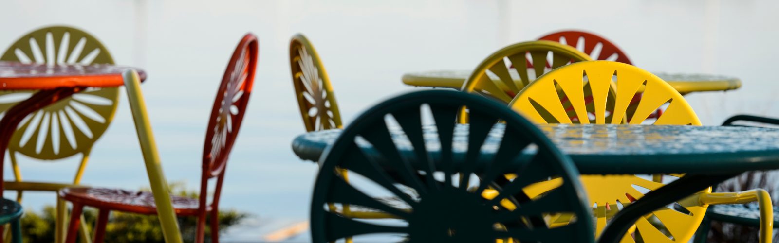 Green, yellow and orange chairs and tables on a terrace with an open lake and boats in the background.